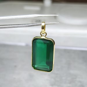 May include: A rectangular emerald-green gemstone pendant with a gold-colored frame and bail. The pendant is held by tweezers against a blurred background. The gemstone is a deep, transparent green.