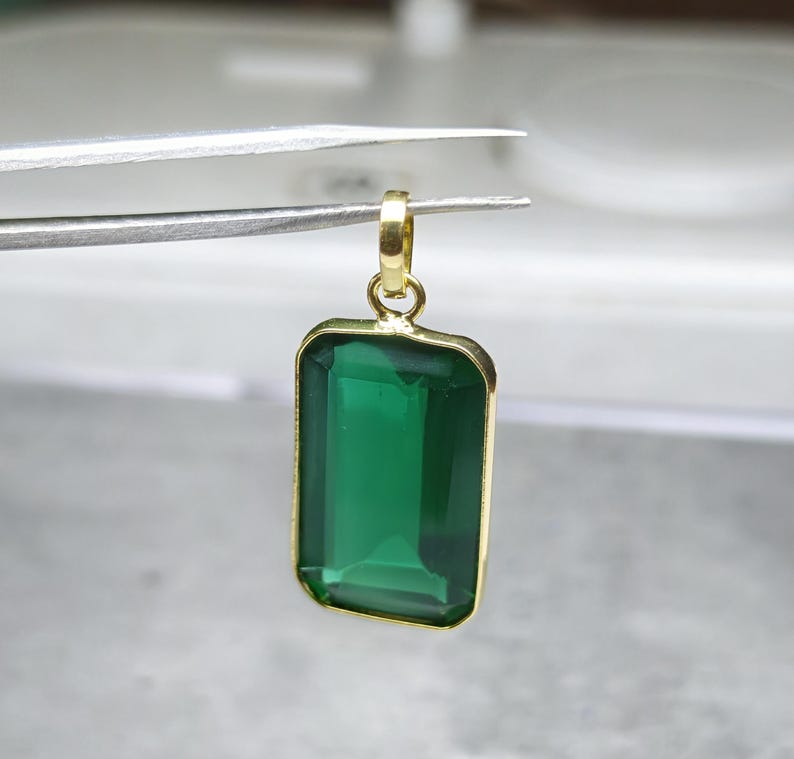 May include: A rectangular emerald-green gemstone pendant with a gold-colored frame and bail. The pendant is held by tweezers against a blurred background. The gemstone is a deep, transparent green.