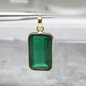 May include: A rectangular green gemstone pendant with a gold-colored frame and bail. The emerald-cut stone is a deep, vibrant green. The pendant is held by tweezers, showcasing its size and design. A piece of jewelry.