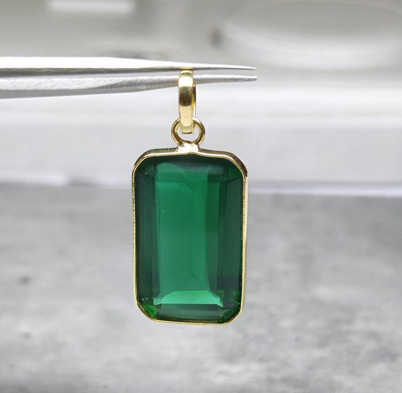 May include: A rectangular green gemstone pendant with a gold-colored frame and bail. The emerald-cut stone is a deep, vibrant green. The pendant is held by tweezers, showcasing its size and design. A piece of jewelry.