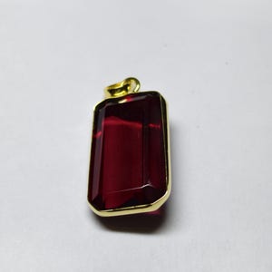 May include: A rectangular pendant featuring a deep red gemstone within a gold-coloured frame. The gemstone has a faceted surface, and the pendant has a small loop at the top for a chain.