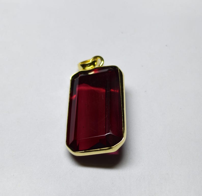 May include: A rectangular pendant featuring a deep red gemstone within a gold-coloured frame. The gemstone has a faceted surface, and the pendant has a small loop at the top for a chain.