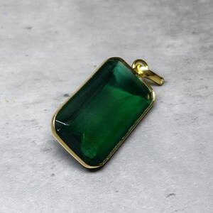 May include: A rectangular pendant with a large, emerald-green gemstone set in a gold-colored metal frame. The pendant has a loop at the top for a chain. The gemstone is faceted, catching the light.