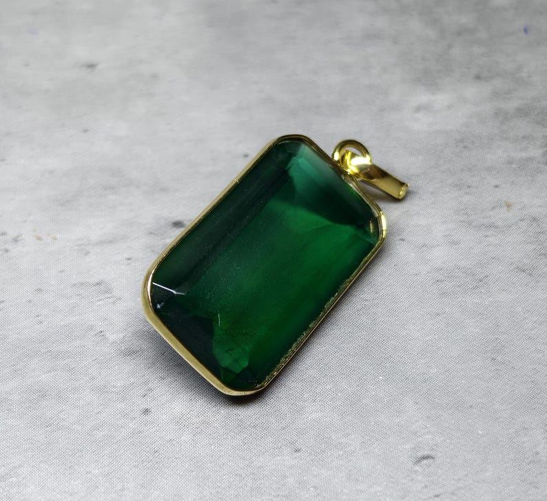 May include: A rectangular pendant with a large, emerald-green gemstone set in a gold-colored metal frame. The pendant has a loop at the top for a chain. The gemstone is faceted, catching the light.
