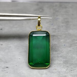 May include: A rectangular, emerald-green gemstone pendant with a gold-colored frame and bail. The pendant is held by tweezers against a neutral background. The gemstone has a faceted cut, reflecting light.