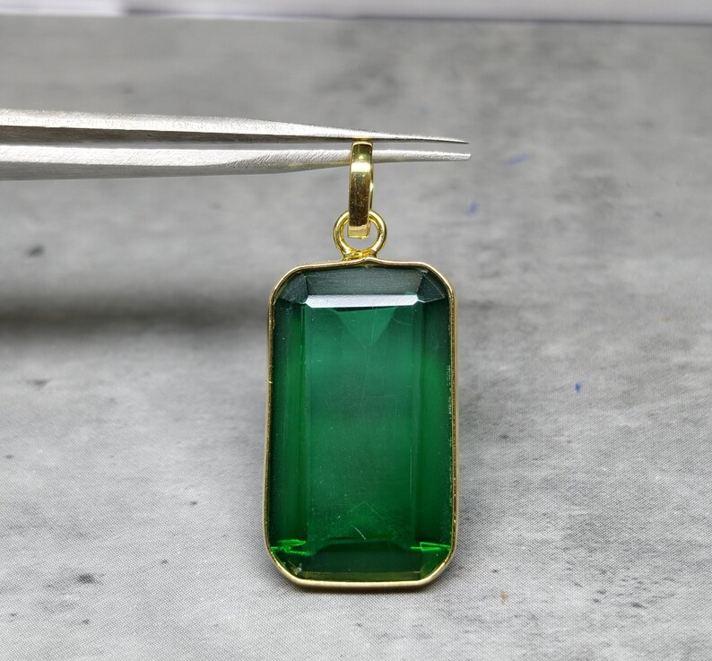 May include: A rectangular, emerald-green gemstone pendant with a gold-colored frame and bail. The pendant is held by tweezers against a neutral background. The gemstone has a faceted cut, reflecting light.