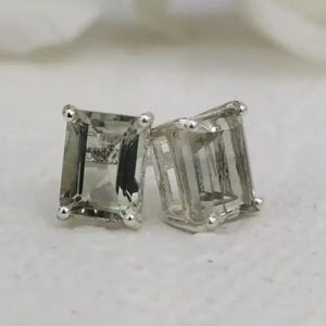 May include: A pair of stud earrings featuring rectangular, emerald-cut light green gemstones set in four-prong silver metal settings. The stones are translucent with visible facets that reflect light, showcasing a pale, cool-toned green hue.