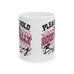 May include: White ceramic mug with the text "I'm holding on mentally, buffering" in black and pink lettering. The mug features a pink and white checkered pattern with daisy accents. The mug is a standard size.