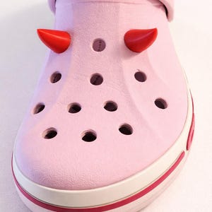 Peut inclure: Un sabot Crocs rose clair avec des cornes de diable rouges attachées. La chaussure a une bande blanche autour de la semelle et une bande rose foncé en bas. Le mot "CROCS" est imprimé sur la bride.