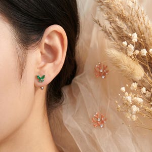 May include: A close-up of a person's ear adorned with a green butterfly earring. The butterfly earring is embellished with small green stones. Two coral-shaped earrings with small white stones are also visible.