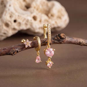 May include: A collection of gold-colored earrings featuring pink gemstones. The set includes a flower-shaped stud, a teardrop-shaped hoop, and a dangling earring with round and heart-shaped stones. The earrings are displayed on a brown branch.
