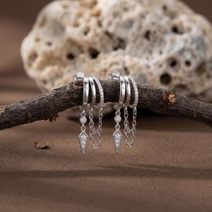 Op de afbeelding: Een paar zilveren oorringen met meerdere banden, versierd met kleine, heldere edelstenen. Elke oorring heeft een bungelende ketting met een ruitvormige hanger. De oorbellen worden gepresenteerd op een kleine tak tegen een neutrale achtergrond.