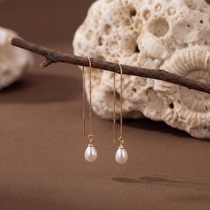 Puede incluir: Un par de pendientes colgantes dorados con perlas blancas en forma de lágrima. Los pendientes cuelgan de una fina cadena dorada y se exhiben en una rama marrón sobre un fondo marrón.