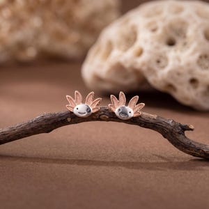 May include: A pair of silver axolotl stud earrings with rose gold accents. The earrings are set on a brown branch against a blurred background. The axolotls have smiling faces and detailed gills.