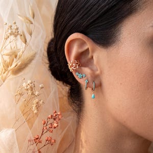 May include: A collection of gold-toned earrings adorns an ear. The earrings include a coral-shaped ear cuff, a cluster of turquoise studs, and small hoop earrings with turquoise accents, one with a turquoise teardrop charm. The jewelry is set against a soft, neutral background.