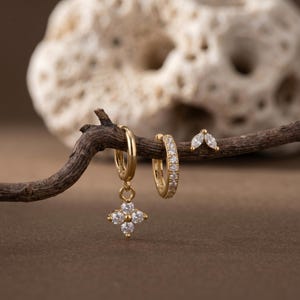 May include: A collection of gold earrings displayed on a small branch. The earrings include a pair of small hoop earrings with clear stones, a pair of hoop earrings with a dangling four-petal flower design, and a pair of stud earrings with a marquise shape.