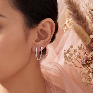 May include: Silver earrings with a chain detail. One earring is a small hoop with clear stones, the other a single clear stone stud. A fine chain connects the two earrings. The design is elegant and suitable for everyday wear or special occasions.