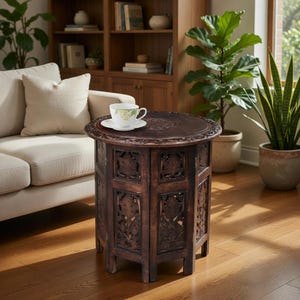 May include: A dark brown, intricately carved wooden side table with a round top and hexagonal base. The table features floral and geometric patterns. A white teacup and saucer sit on the table's surface. The table is approximately 24 inches tall.