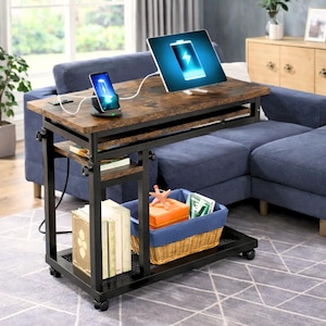 May include: A brown and black rolling side table with a tablet and phone charging on top. The table has a rustic wood-look surface and a black metal frame. A blue sofa is in the background. The table has a shelf and a basket for storage.