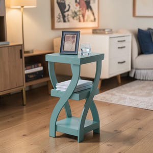 Puede incluir: Una mesa auxiliar turquesa con un diseño único de reloj de arena. La mesa tiene tres niveles, con una parte superior cuadrada y dos estantes inferiores. Una foto enmarcada y un vaso de agua están encima. La mesa mide unos 61 cm de alto.