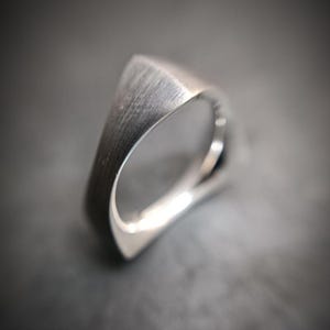 May include: A silver-toned ring with a triangular top section featuring a brushed, textured finish. The band has a smooth, polished interior surface and a distinct geometric shape that tapers toward the bottom, creating a clean and angular profile.