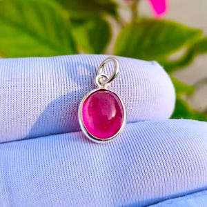 Op de afbeelding: Een kleine ovale hanger met een gladde, gepolijste felroze edelsteen in een eenvoudige zilverkleurige metalen zetting. De hanger is voorzien van een ronde zilverkleurige ring aan de bovenzijde voor bevestiging aan een ketting of een koord.