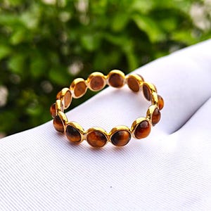 May include: A gold ring featuring a series of oval-shaped, brown tiger's eye stones. The ring is set on a white surface, with a blurred green background.
