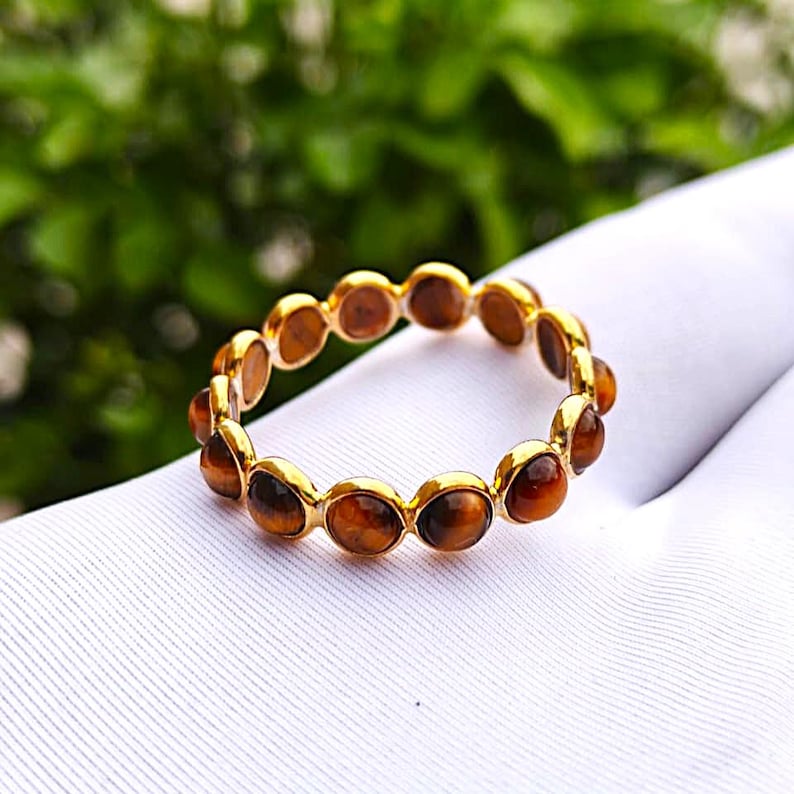 May include: A gold ring featuring a series of oval-shaped, brown tiger's eye stones. The ring is set on a white surface, with a blurred green background.