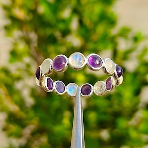 Op de afbeelding: Een zilveren ring met een afwisselend patroon van ronde, in een zetting geplaatste paarse amethist en iriserende witte maansteen cabochons. De stenen zijn gerangschikt in een doorlopende cirkelvormige band, vastgehouden door een metalen hulpmiddel.
