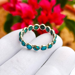 Op de afbeelding: Een zilveren ring met ovale turkooizen stenen rond de band. De ring wordt vastgehouden door een gehandschoende hand, met rode bloemen op de achtergrond. De turkooizen stenen zijn blauwgroen met een bruine matrix.
