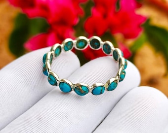 May include: A silver ring with oval turquoise stones set around the band. The ring is held by a gloved hand, with red flowers in the background. The turquoise stones have a blue-green color with brown matrix.