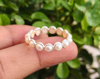 May include: A silver ring featuring a continuous row of small, round, white pearls. The ring is set against a blurred background of green foliage. The pearls are evenly spaced around the band.