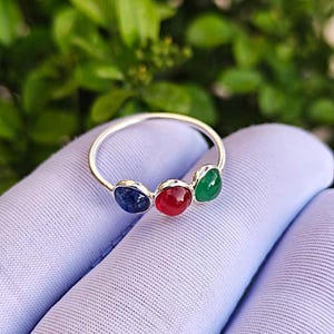 Può includere: Un anello d'argento con tre pietre preziose: un ovale blu, un tondo rosso e una goccia verde. L'anello è posto su uno sfondo di tessuto viola chiaro, con fogliame verde sullo sfondo sfocato.