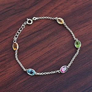 Peut inclure: Un bracelet en argent avec une chaîne délicate. Il est orné de six pierres précieuses ovales de couleurs variées : jaune, vert, bleu et rose. Le bracelet est présenté sur une surface en bois sombre.