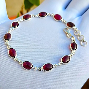Könnte beinhalten: Ein silbernes Kettenarmband mit zehn ovalen, tiefroten Granat-Edelsteinen, die in polierte Metallfassungen eingefasst sind. Das Armband verfügt über einen Karabinerverschluss und eine kurze Verlängerungskette mit drei runden Gliedern am Ende.