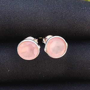 Peut inclure: Une paire de boucles d'oreilles rondes avec une pierre précieuse rose clair à facettes sertie dans un métal argenté. Les boucles d'oreilles sont présentées sur une surface sombre et texturée.