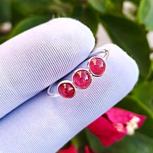 May include: A silver ring featuring three round, vibrant red gemstones. The gemstones are set in a row, creating a unique and eye-catching design. The ring is displayed on a light blue fabric, highlighting the ring's color and shine.