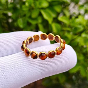 May include: A gold-colored ring with a series of oval, amber-colored gemstones. The ring is held against a white fabric background, with green foliage visible in the background. The gemstones are set in individual gold bezels.