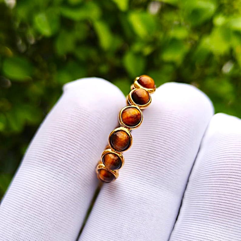 May include: A gold ring featuring a series of round, polished tiger's eye stones. The stones are a warm brown color with golden striations, set in a gold band. The ring is displayed on a white surface against a blurred green background.