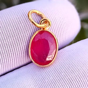 Peut inclure: Un pendentif doré avec une pierre précieuse rouge rubis ovale et facettée. Le pendentif a une boucle pour une chaîne. La pierre est sertie dans une lunette dorée. Il s'agit probablement d'un bijou.