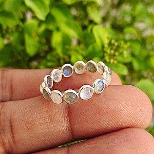 May include: A silver ring with a band of small, round gemstones in white, blue, and grey. The gemstones are set closely together, forming a continuous circle. The ring is displayed against a backdrop of green leaves.