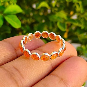 Op de afbeelding: Een zilveren ring met een doorlopende band van ovale, oranje edelstenen. Het ontwerp van de ring wordt benadrukt tegen een vage groene achtergrond. De kleur van de edelstenen is een opvallend kenmerk van de ring.