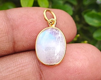 May include: An oval-shaped pendant featuring a translucent, iridescent moonstone set in a polished gold-toned metal bezel. The pendant includes a small, circular gold-toned jump ring at the top for attachment to a chain or cord.