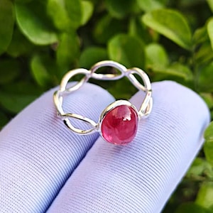 Op de afbeelding: Een zilveren ring met een ovale, levendige rode edelsteen. De band van de ring heeft een open, verweven ontwerp. De edelsteen is gezet in een zilveren bezel, wat een opvallend contrast creëert. De ring wordt getoond tegen een zachte, lichtblauwe achtergrond.
