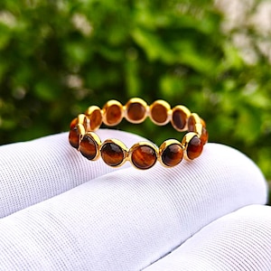May include: A gold-colored ring featuring a series of round, tiger's eye gemstones. The gemstones are a rich brown color with golden streaks. The ring is set on a white surface with a blurred green background.