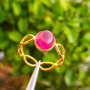 Könnte beinhalten: Ein goldener Ring mit einem großen, ovalen, rosa Edelstein. Das Band des Rings hat ein durchbrochenes Design. Der Edelstein ist der Mittelpunkt, vor einem verschwommenen grünen Hintergrund.