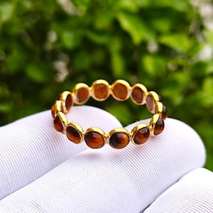 May include: A gold-colored ring with a series of oval-shaped, brown gemstones set around the band. The ring is held against a white fabric background, with a blurred green foliage backdrop.