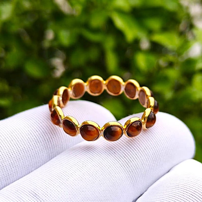May include: A gold-colored ring with a series of oval-shaped, brown gemstones set around the band. The ring is held against a white fabric background, with a blurred green foliage backdrop.