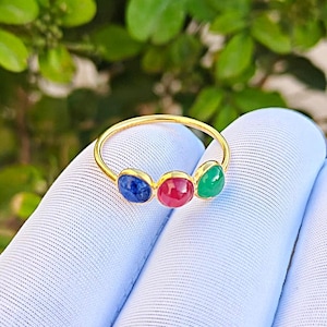 May include: A gold ring featuring three gemstones: a blue sapphire, a red ruby, and a green emerald. The gemstones are set in a row along the ring's band. The ring is displayed against a light blue fabric with a blurred green background.