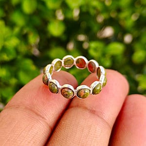 Op de afbeelding: Een zilveren ring met een band van ovaalvormige, groen en rode edelstenen. De ring wordt in een hand gehouden tegen een vage groene achtergrond. De edelstenen zijn gezet in individuele zilveren zettingen.